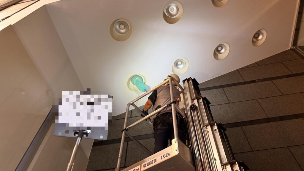 高所作業台に乗った作業員が、天井のダウンライトをグリーンのブラシで清掃している様子。「1F 作業中」の表示板が手前に立っている