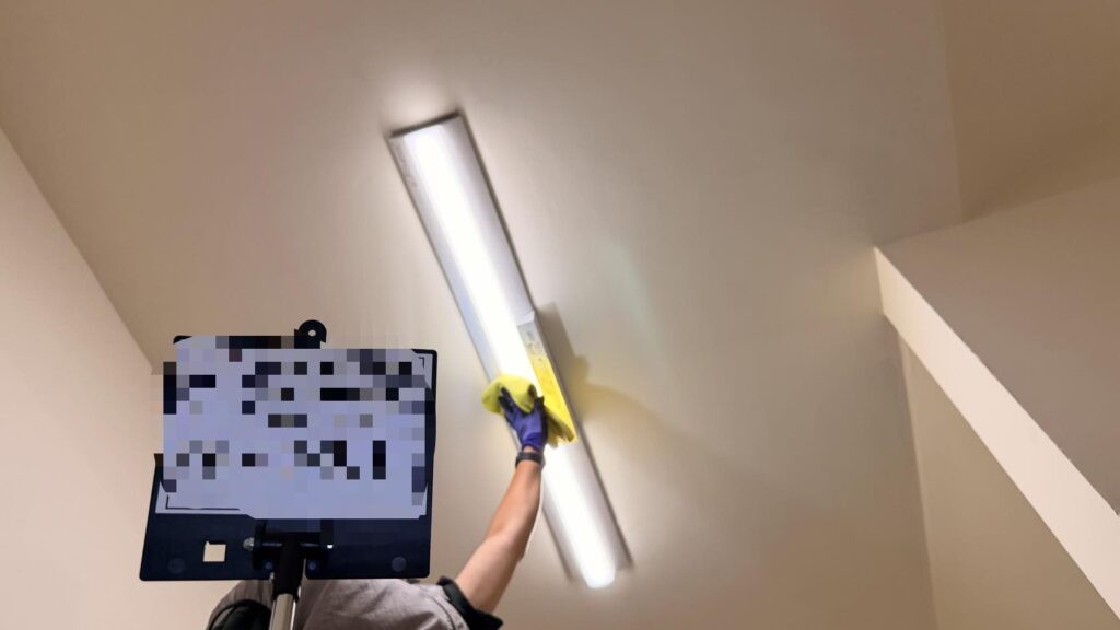 階段の高い位置にある細長い照明器具を、作業員が黄色いクロスで拭き上げている様子