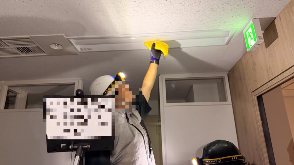 ヘルメットとヘッドライトを着けた作業員が、天井の照明器具を黄色いクロスで拭き上げている様子