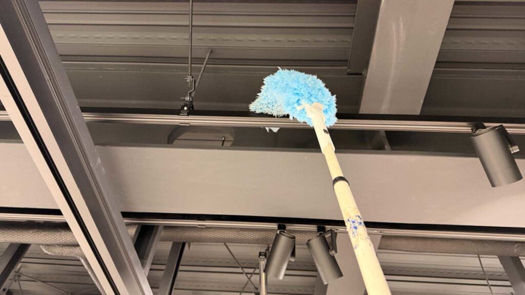 高所の梁や照明器具まわりのホコリを、作業員が青いマイクロファイバー製の伸縮ダスターで除塵している様子