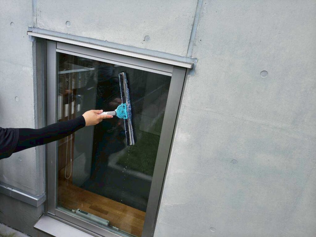打ちっぱなしコンクリートの壁にはめ込まれた窓ガラスを、作業員がスクイジーで清掃している様子